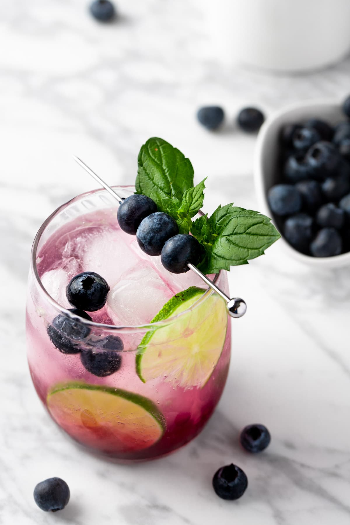 A blueberry mojito mocktail garnished with blueberries, on a white marble table.