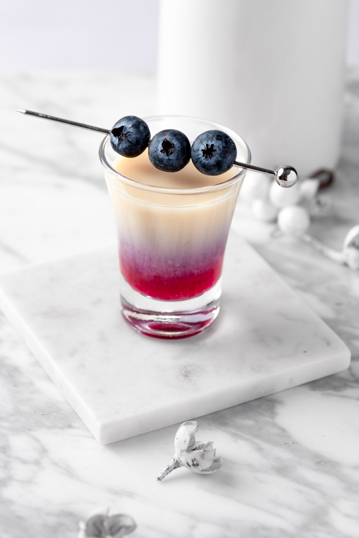 A blueberry muffin shot garnished with 3 blueberries on a white table.