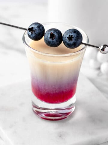 A blueberry muffin shot garnished with 3 blueberries on a white table.
