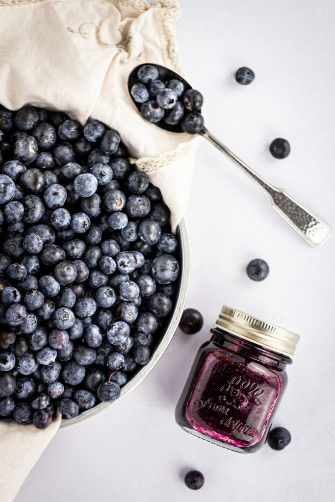 Overhead photo of a metal tin bowl filled with fresh blueberries, a jar of blueberry syrup and a spoon holding a few blueberries.