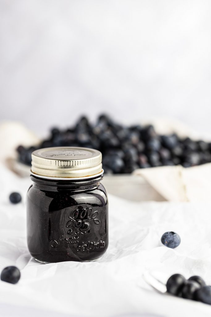 A glass jar of blueberry syrup with a bowl filled with fresh blueberries in the background.