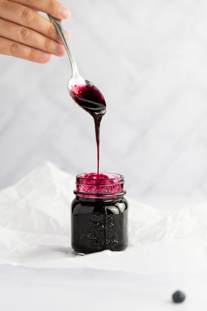 A hand holding a metal spoon above the open jar of blueberry syrup, drizzling the syrup into the jar.