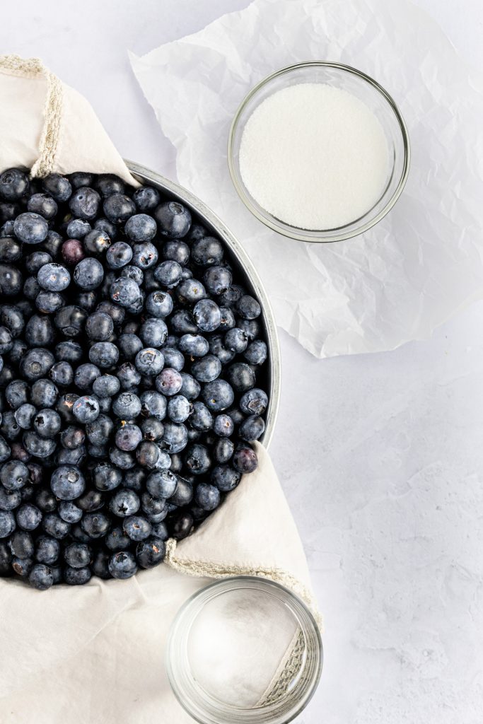 Overhead photo of fresh blueberries on a beige napkin in a metal tin, a glass of water and a small glass bowl of granulated sugar.