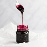 A hand holding a metal spoon above the open jar of blueberry syrup, drizzling the syrup into the jar.