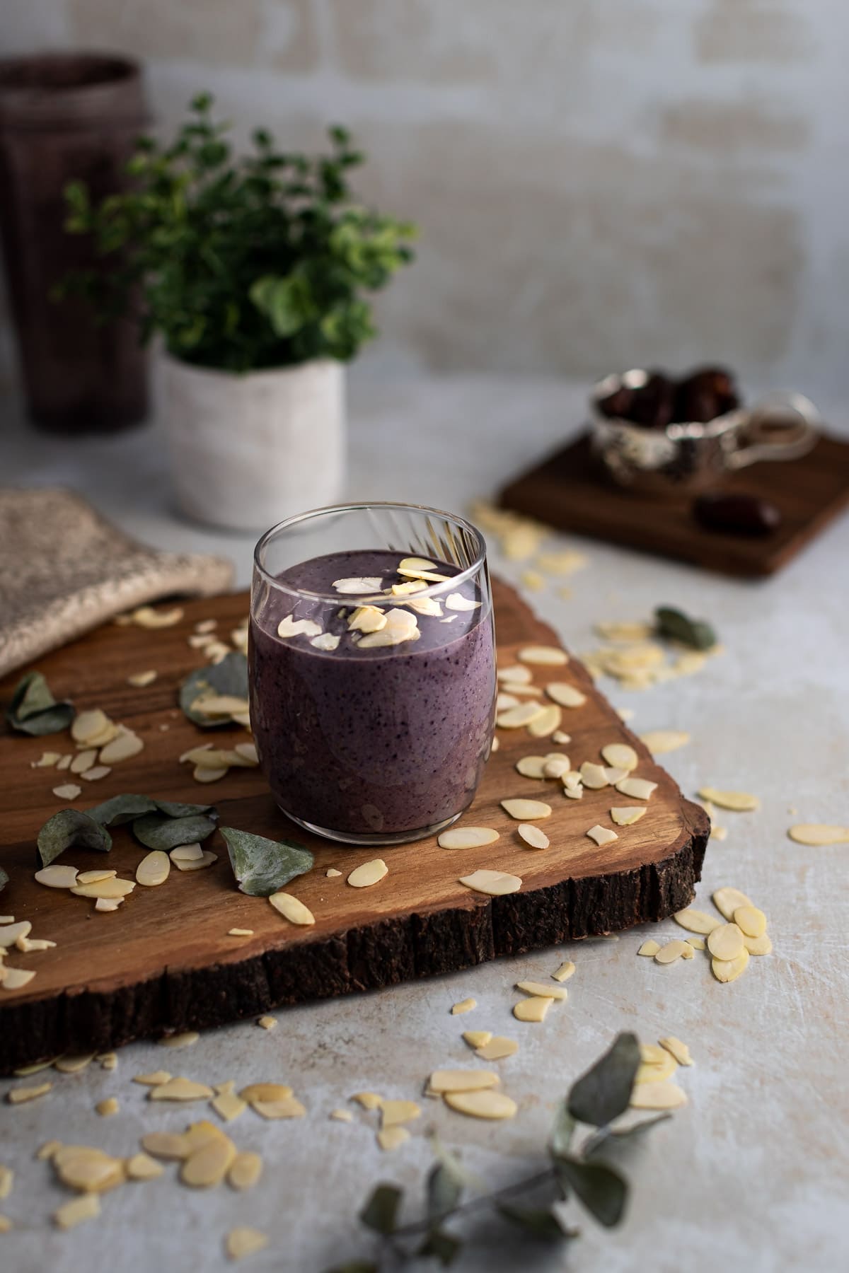 Glass of tahini smoothie, topped with sliced almonds, on a wooden board, with a green plant in the background.