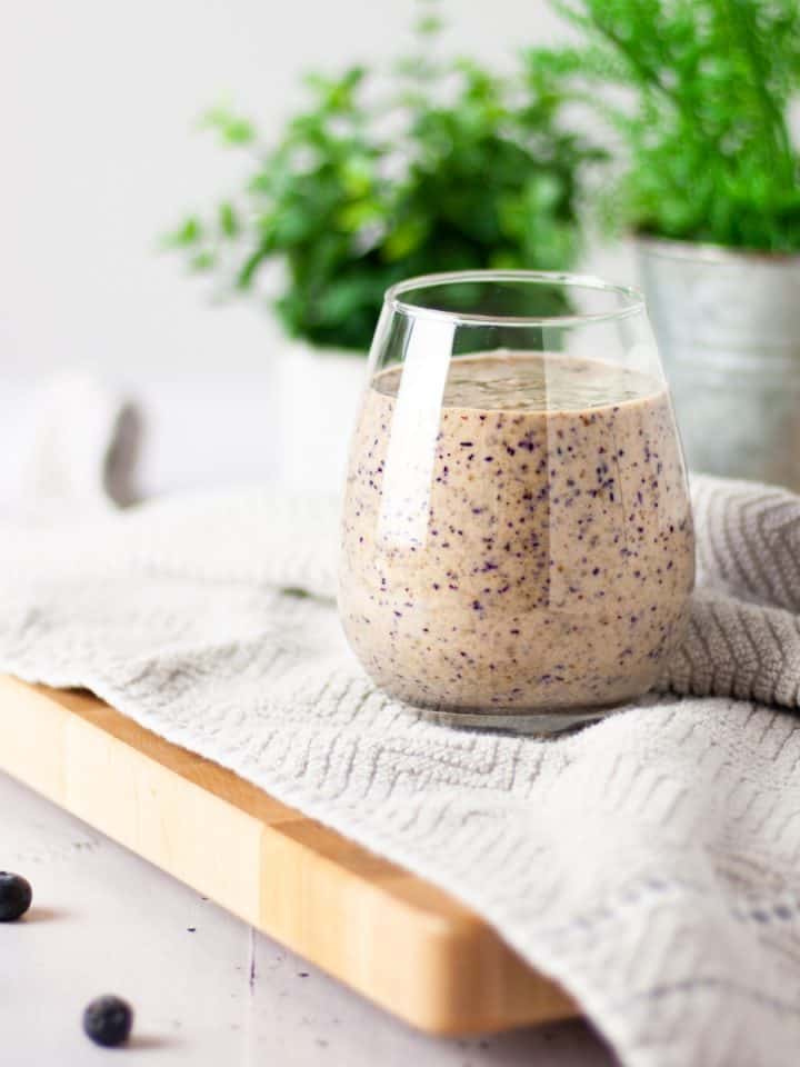 A glass of blueberry tahini smoothie on a light brown hand towel with a plant in the background and scattered blueberries in the foreground