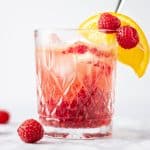 Eye-level view of a glass of bourbon raspberry cocktail with a couple raspberries and an orange slice as garnish.