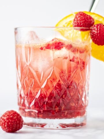 Eye-level view of a glass of bourbon raspberry cocktail with a couple raspberries and an orange slice as garnish.