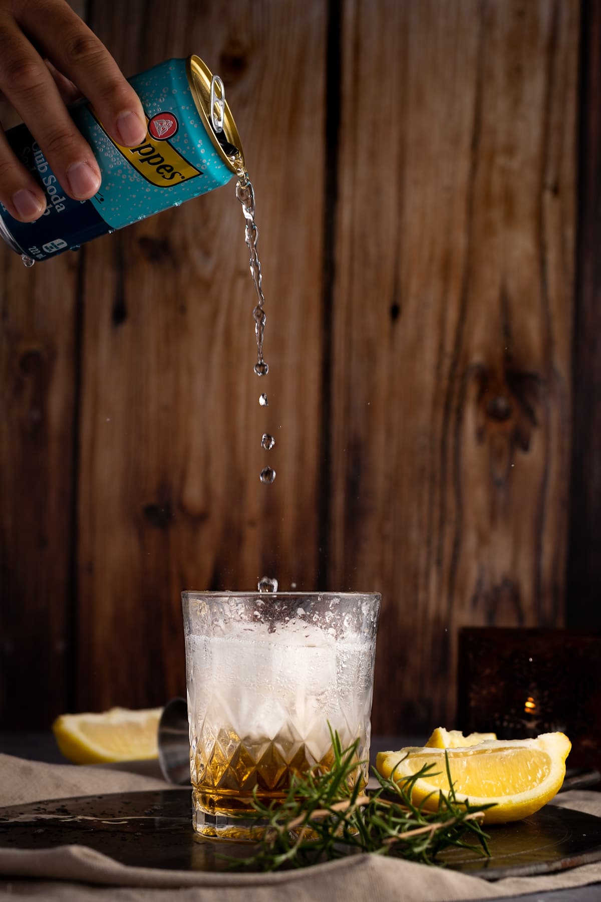 Club soda being poured into the cocktail, sitting on a dark metal plate, next to rosemary and lemon slices, with a dark wood background.