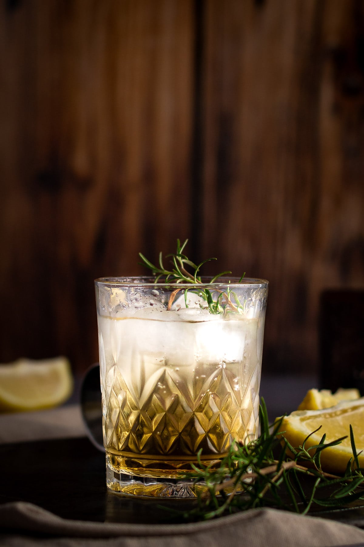 A golden bourbon rosemary cocktail, garnished with rosemary, sitting on a dark metal tray, next to lemon slices with a wooden background.