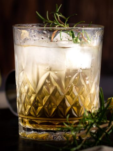 A bourbon rosemary cocktail, garnished with fresh rosemary, with a dark wooden background.