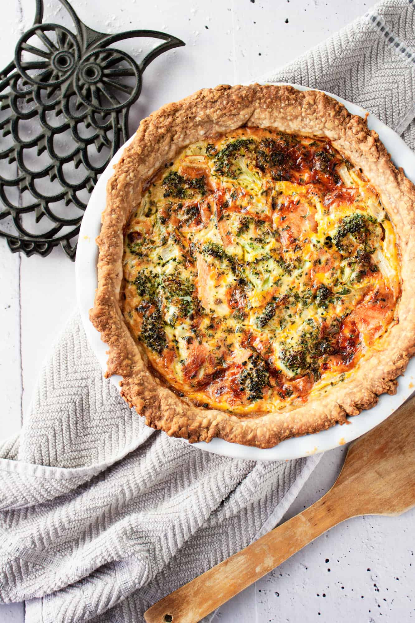 Overhead view of broccoli and smoked salmon quiche fresh out of the oven, on a grey hand towel, next to an owl trivet