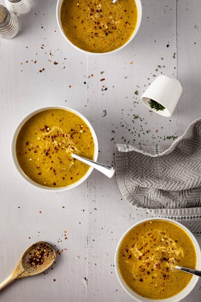 Overhead view of 3 bowls of sweet potato and broccoli soup, a wooden spoon holding red pepper flakes and spilt cup of dried parsley
