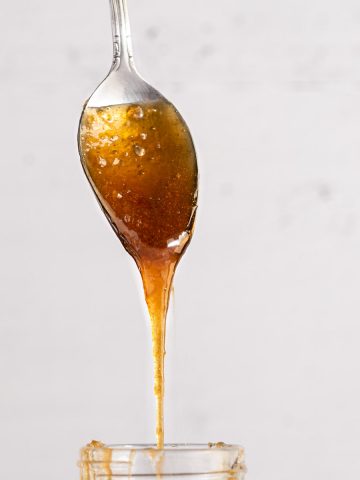 Up close of spoon coated in brown sugar syrup, dripping down into a glass container, on a white background.