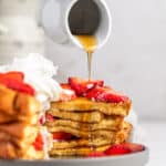 Maple syrup being poured over a stack of buttermilk french toast, topped with fresh strawberries.