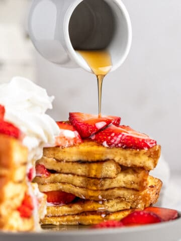 Maple syrup being poured over a stack of buttermilk french toast, topped with fresh strawberries.
