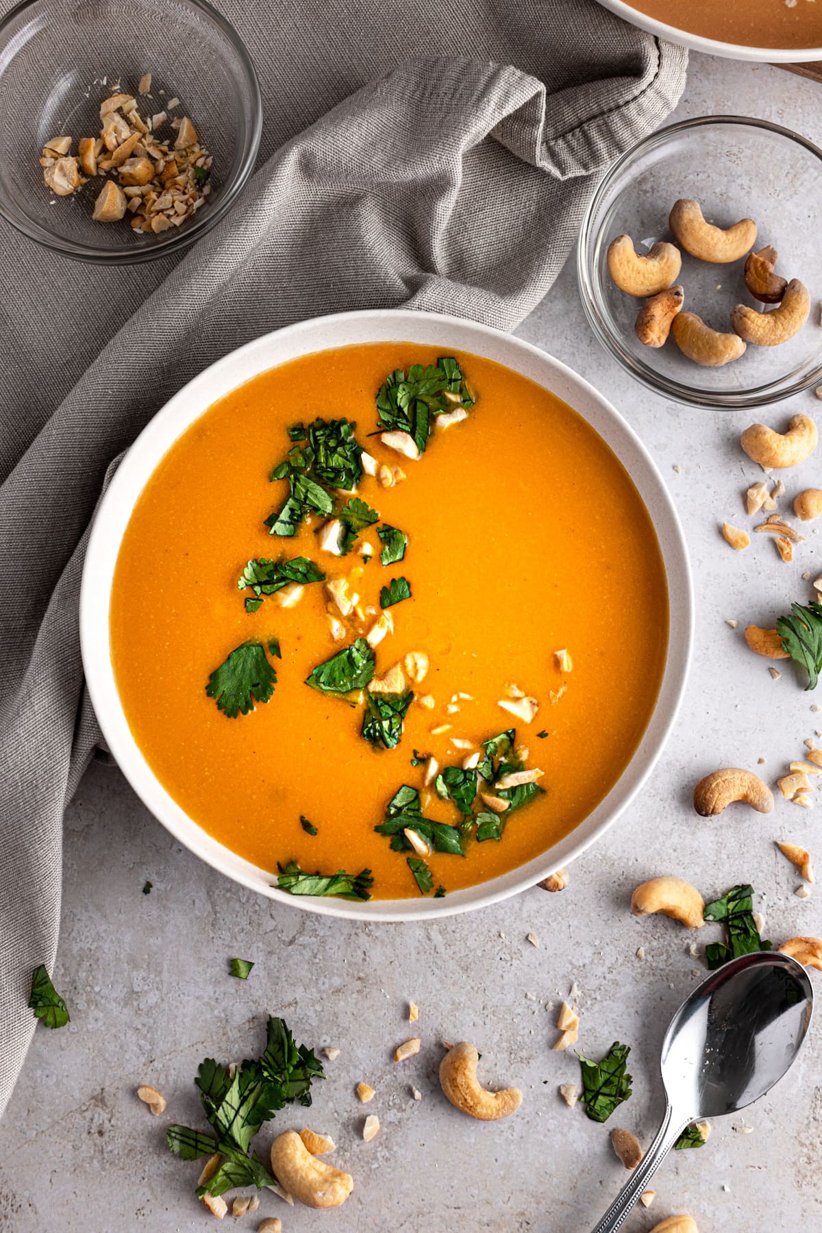 Cashew carrot ginger soup garnished with cilantro and crushed cashews, on a grey table, next to a grey napkin.