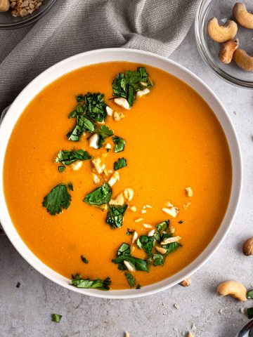 Cashew carrot ginger soup garnished with cilantro and crushed cashews, on a grey table, next to a grey napkin.