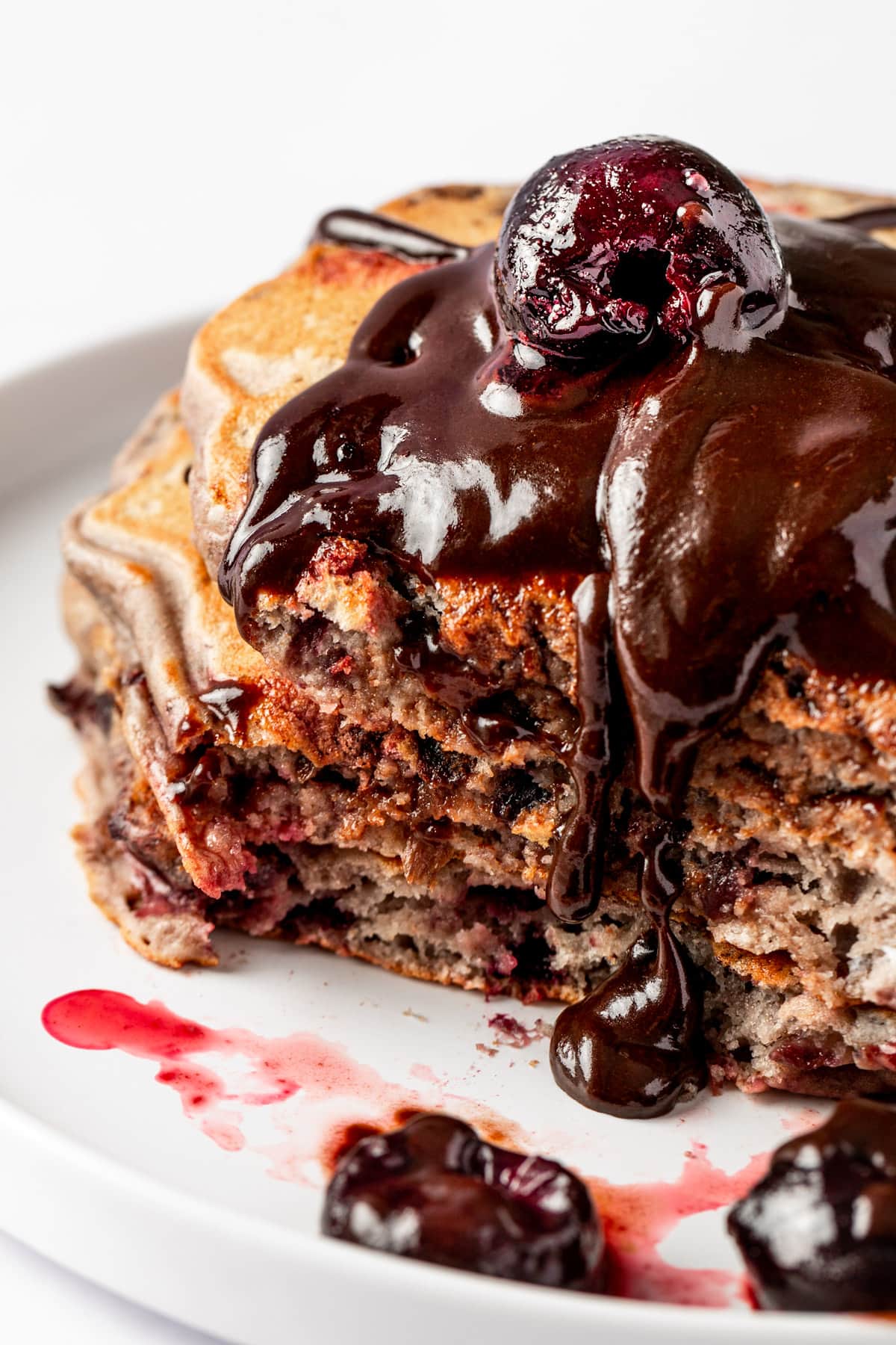 A stack of cherry pancakes with a bite missing, topped with chocolate sauce and a fresh cherry.