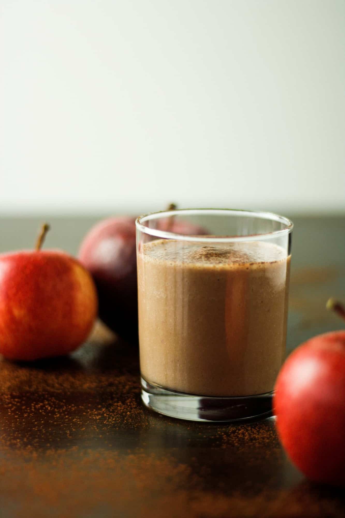 A glass of chocolate apple smoothie on a wooden table next to three apples.