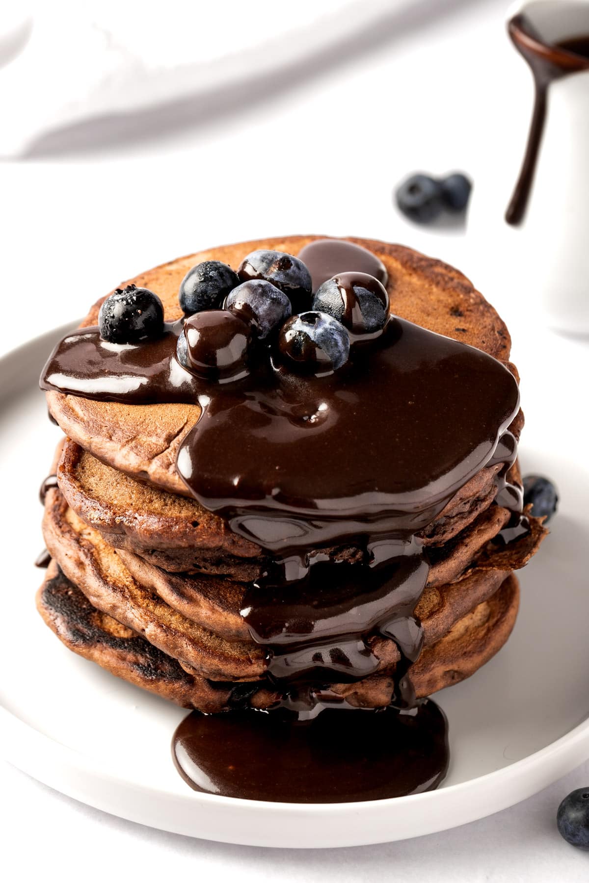 A stack of chocolate chai spiced pancakes drizzled in chocolate pancake sauce, topped with blueberries, on a white plate.