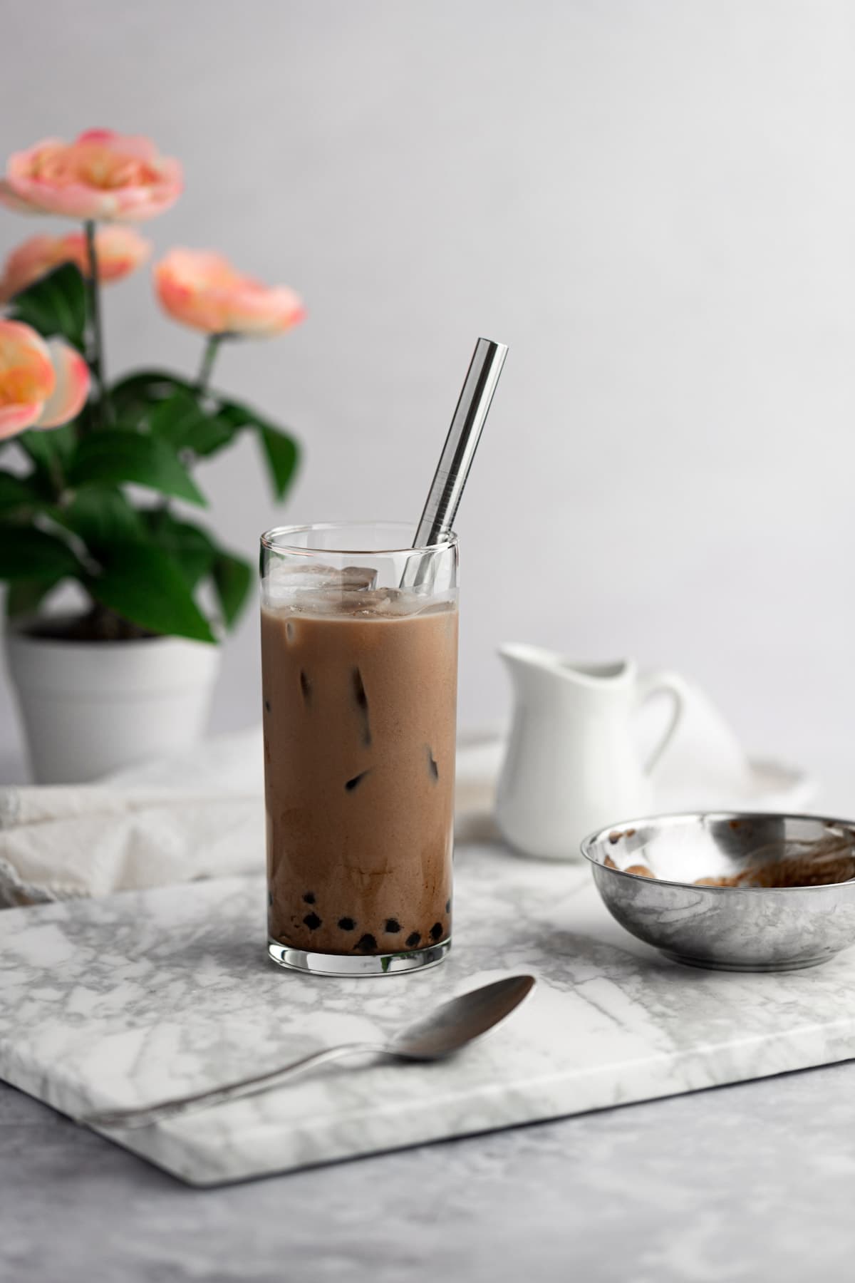 A glass of chocolate milk tea on a grey marble board, next to a metal spoon, with a flower pot in the background.