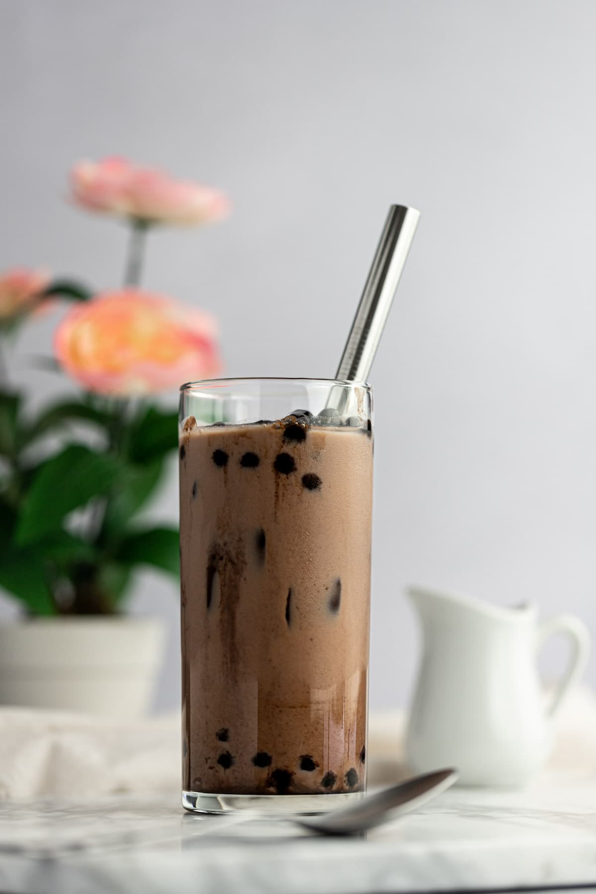 Up close photo of a glass of chocolate milk tea, filled with ice and boba, with a spoon in front of the glass, and a small creamer behind, on a white background.