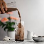 Chocolate milk tea being poured into a glass filled with chocolate soaked boba and ice, pink flowers in the background in front of a white wall.