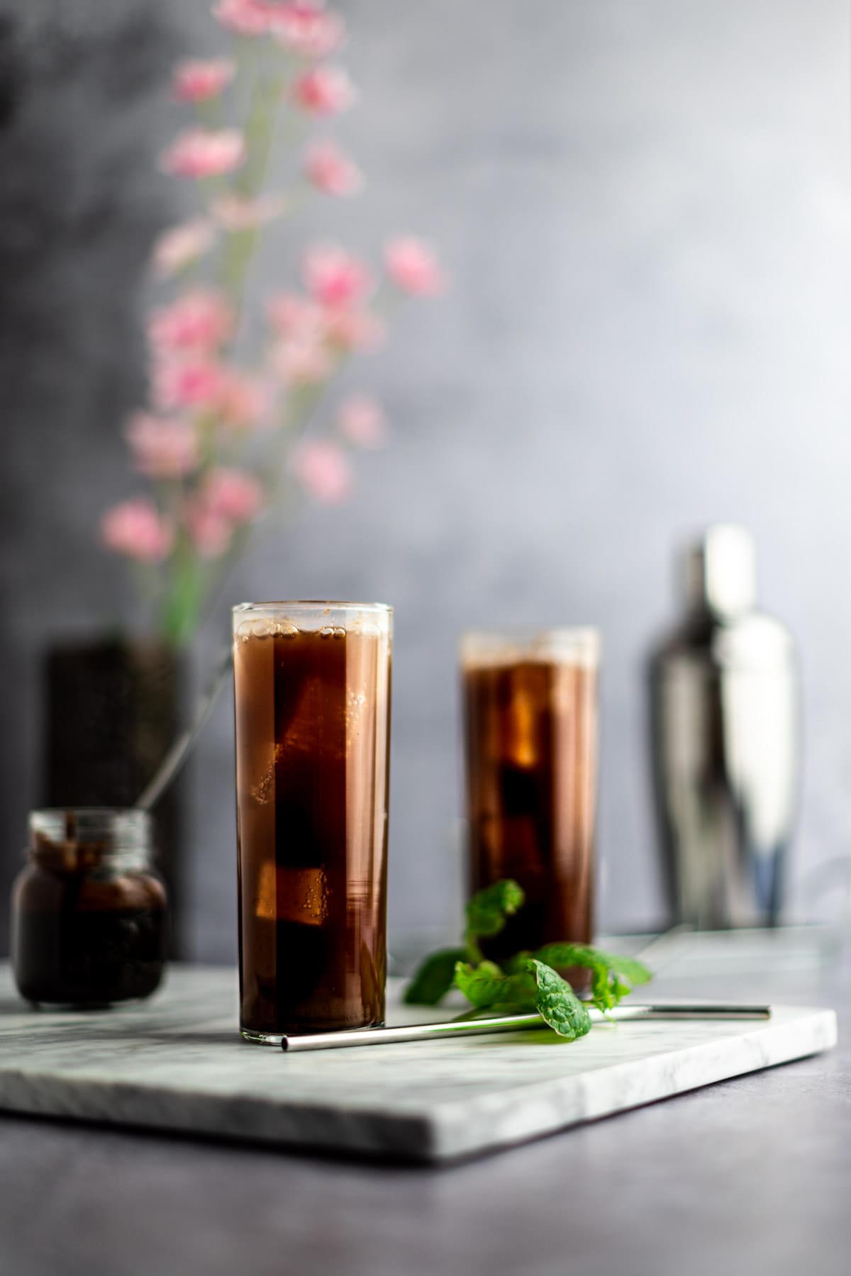 A couple mojitos on a marble board, next to a metal spoon, mint leaves and a jar of chocolate syrup, with flowers and a cocktail shaker in the background.