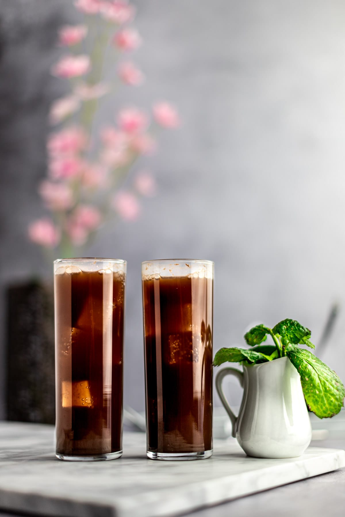 Two chocolate mojitos lined up side by side on a white marble board, next to a small white jug filled with fresh mint leaves.