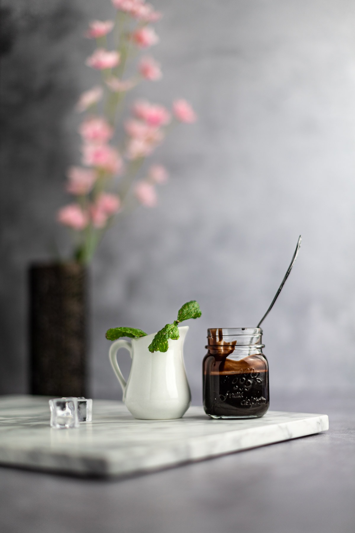 A small white jar filled with mint leaves sitting next to a mason jar filled halfway with chocolate syrup and a spoon sitting in the jar, pink flowers in the background.