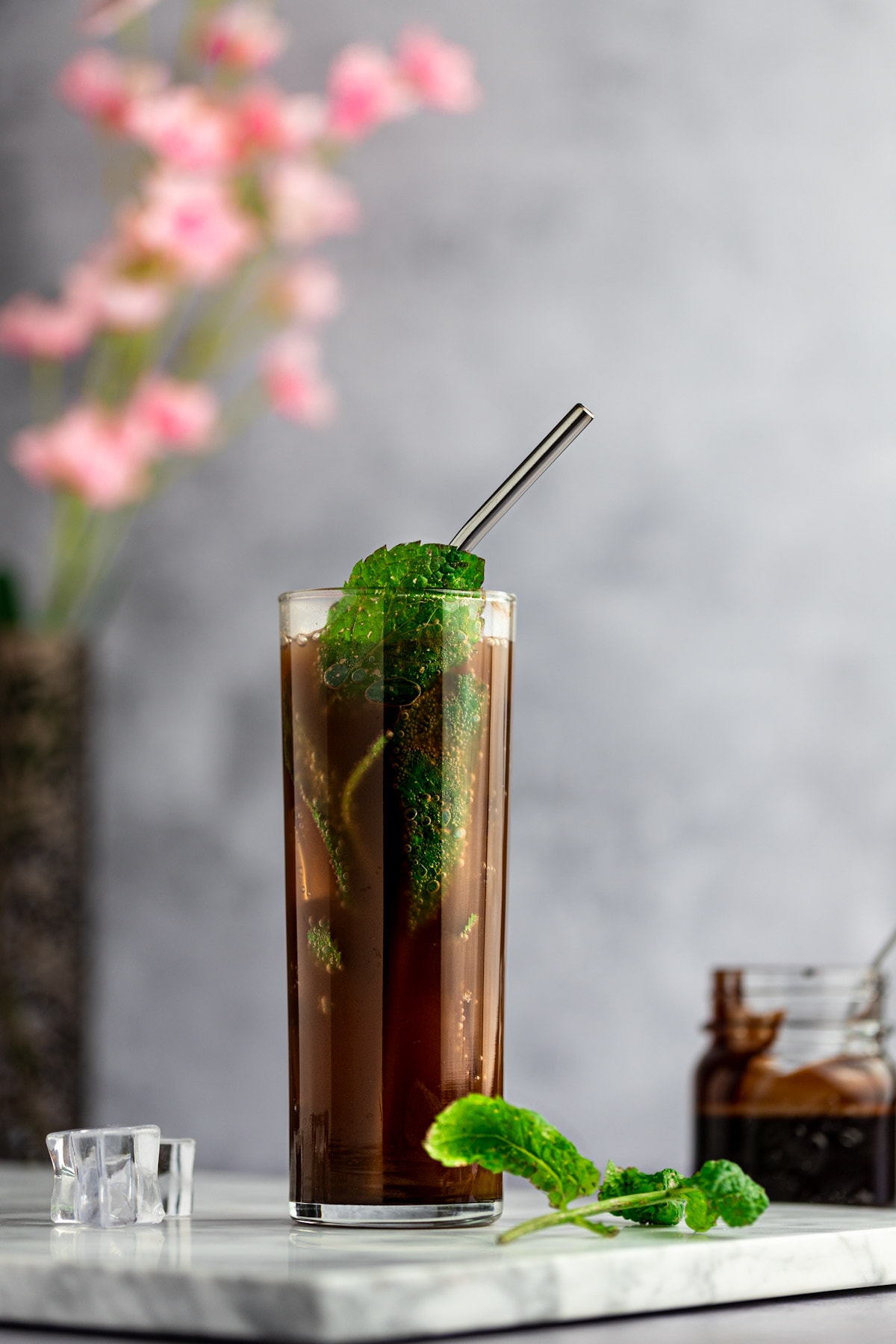 Up close photo of a chocolate mojito, with a metal straw and garnished with fresh mint leaves, on a marble board, next to mint leaves and ice cubes, with pink flowers behind.