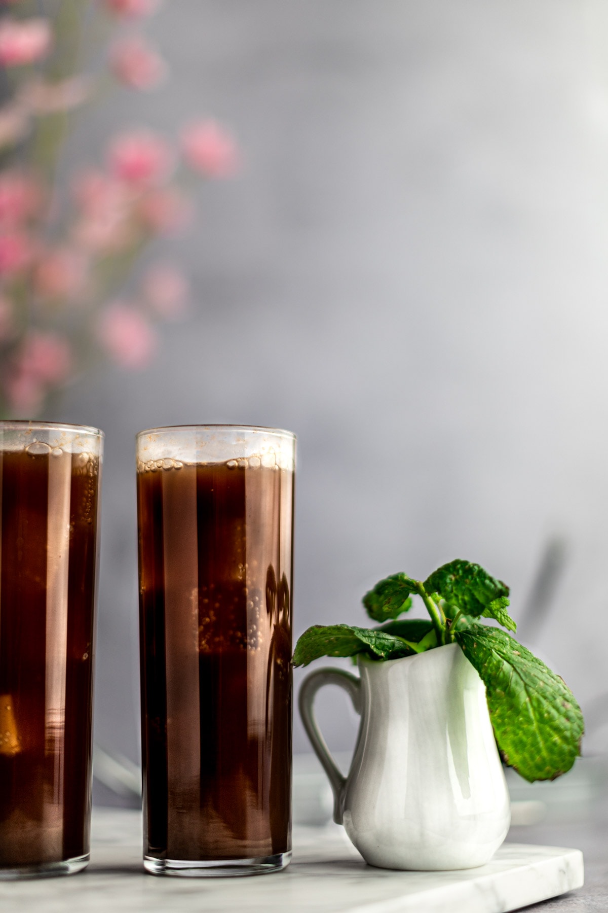 Two glasses of chocolate mojito and a white jar filled with fresh mint lined up beside each other, on a marble board, with flowers in the background.