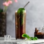 Up close photo of a chocolate mojito, with a metal straw and garnished with fresh mint leaves, on a marble board, next to mint leaves and ice cubes, with pink flowers behind.