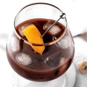 A chocolate old fashioned garnished with an orange peel, on a white marble table.