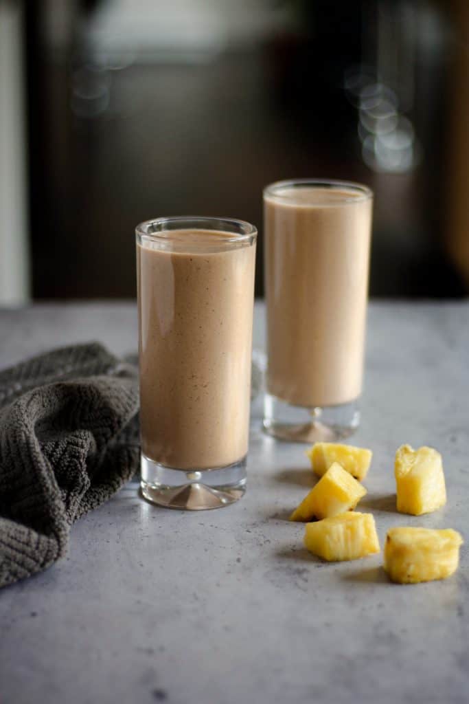 Two tall glasses of chocolate pineapple smoothie on the table with pineapple chunks and a brown hand towel beside them