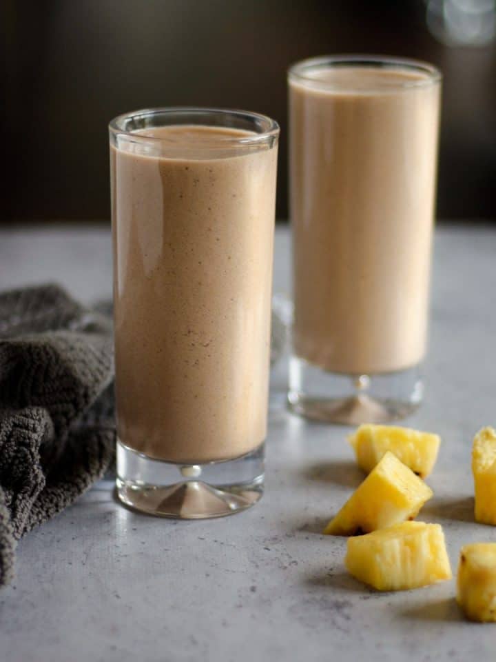 Two tall glasses of chocolate pineapple smoothie on the table with pineapple chunks and a brown hand towel beside them