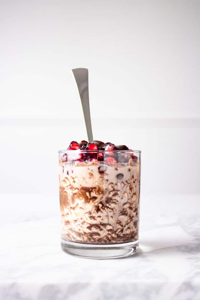 A glass of chocolate pomegranate overnight oats with a silver spoon sticking out