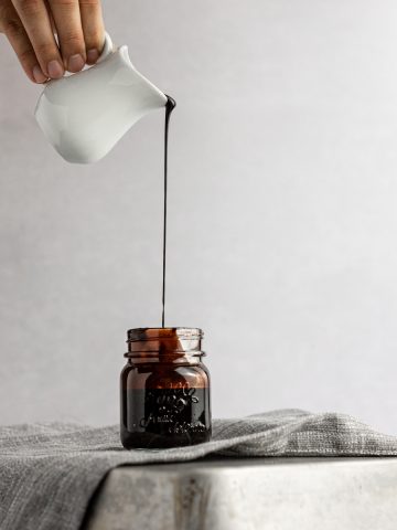 Chocolate simple syrup being poured into a small glass jar, sitting on a grey napkin on a reversed metal tin pan.