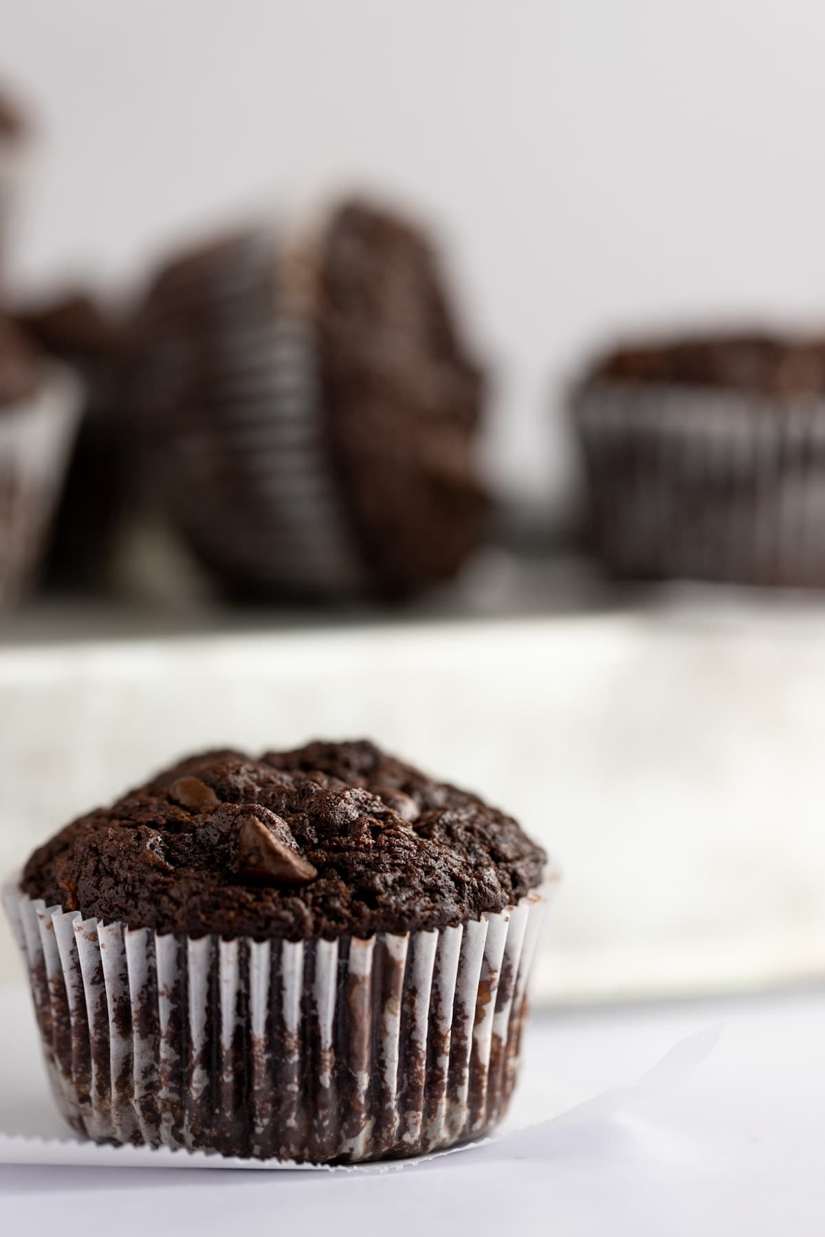 Close up of a sweet potato chocolate chip muffin with its wrapper on, and a tray of muffins out of focus in the background.