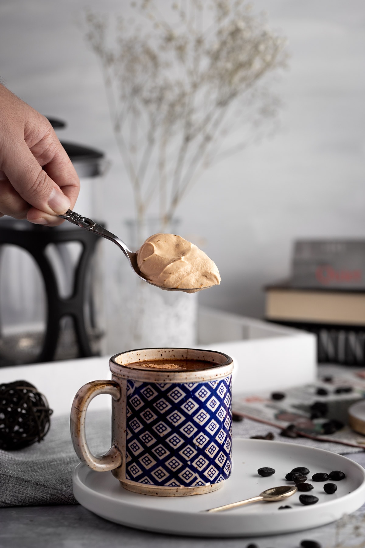 A scoop of coffee whipped cream being held on a metal spoon above a mug of hot chocolate, with a french press, glass vase and books in the background.
