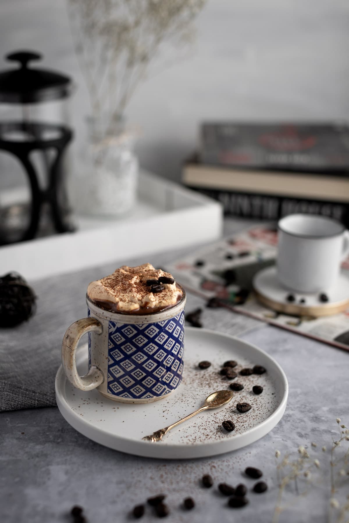 Caffeinated hot chocolate topped with coffee whipped cream in a blue and gray geometric patterned mug, on a white plate with books in the background.