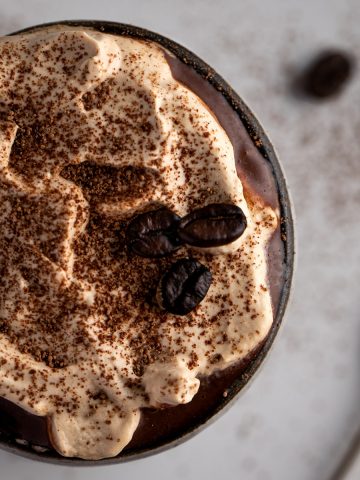 Overhead view of coffee hot chocolate topped with a light brown coffee whipped cream, sprinkle of cocoa powder and 3 coffee beans.