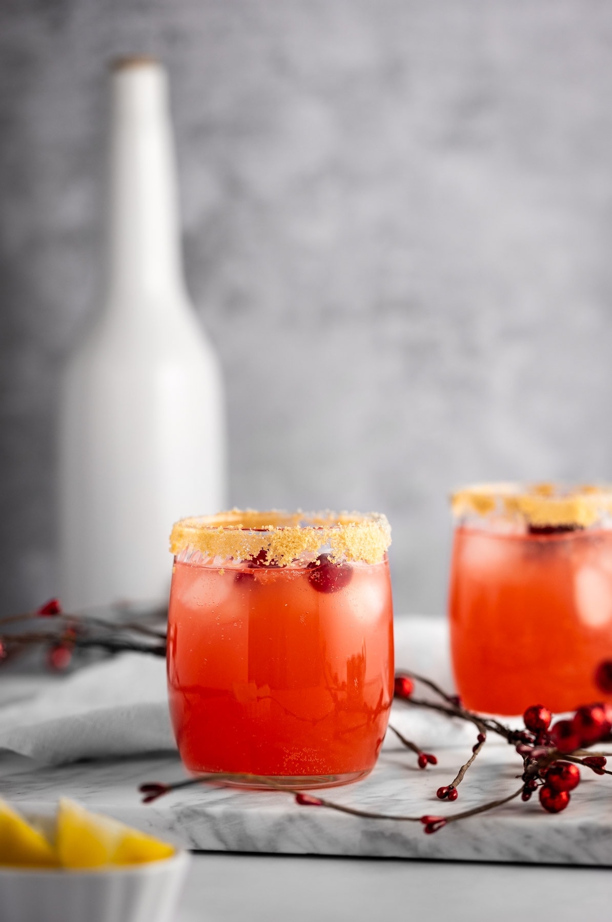 A couple red cranberry drinks on a white marble board, with brown sugar rims, on a grey background.