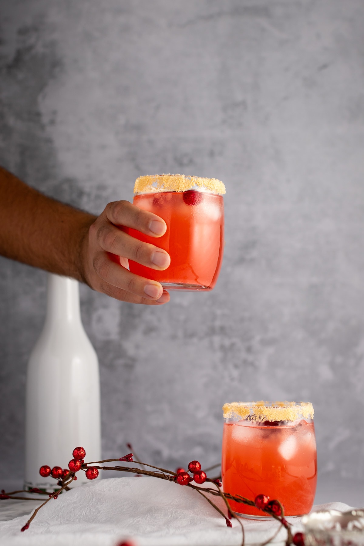 A hand holding a red cranberry mocktail with a brown sugar rim, on a grey, speckled background.
