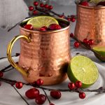 Two cranberry moscow mules in copper mugs, garnished with fresh cranberries and a slice of lime, on a grey background.