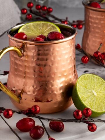 Two cranberry moscow mules in copper mugs, garnished with fresh cranberries and a slice of lime, on a grey background.