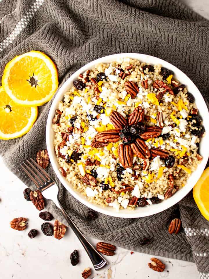 Overhead view of a white bowl of cranberry pecan salad on a grey hand towel, with orange slices, pecans and a fork on the table beside it.