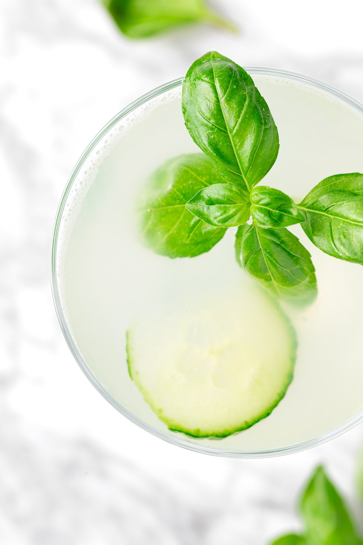 A cucumber basil gimlet garnished with basil, on a white marble table.
