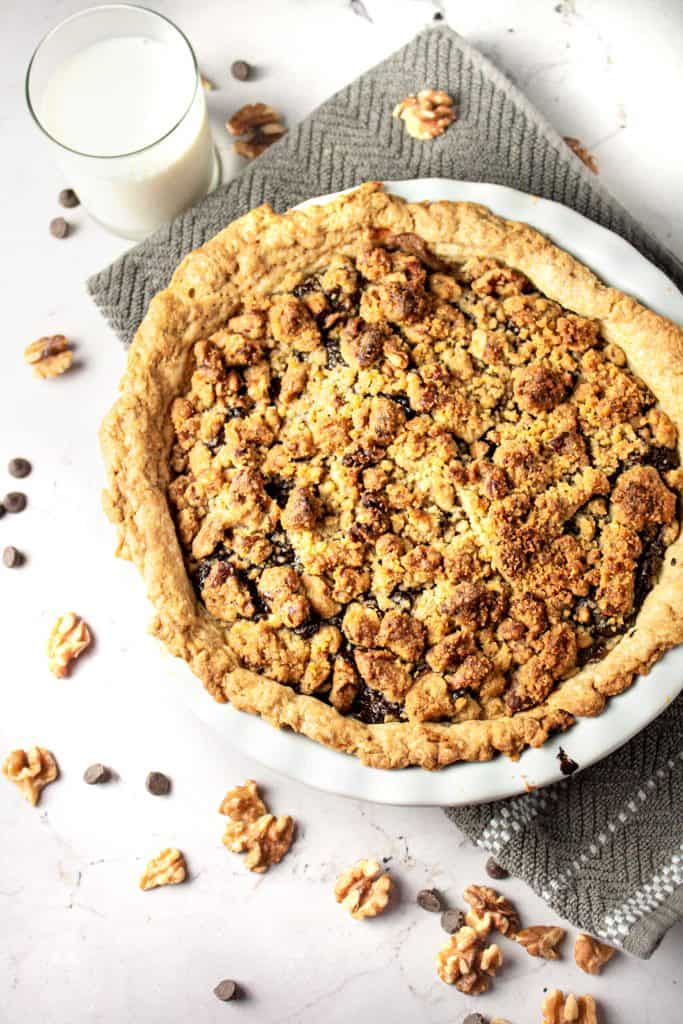 Overhead view of a whole pear chocolate crumble pie next to a glass of milk, on a marble white background with a grey hand towel